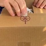 Two hands lifting the tape on a plain cardboard carton stamped with the rune mark.